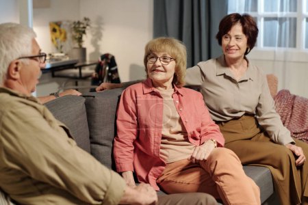 Téléchargez les photos : Groupe de grands-parents heureux en tenue décontractée ayant parlé de leurs intérêts tout en se relaxant sur un canapé confortable et doux dans le salon - en image libre de droit