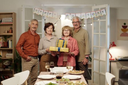 Téléchargez les photos : Groupe de quatre amis aînés heureux avec des boîtes-cadeaux emballés regardant la caméra tout en profitant fête d'anniversaire de l'un d'eux - en image libre de droit