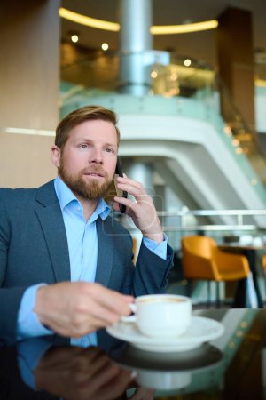 Téléchargez les photos : Jeune homme d'affaires avec smartphone par oreille communiquer avec des partenaires ou consulter ses subordonnés tout en prenant une tasse de café dans un café - en image libre de droit