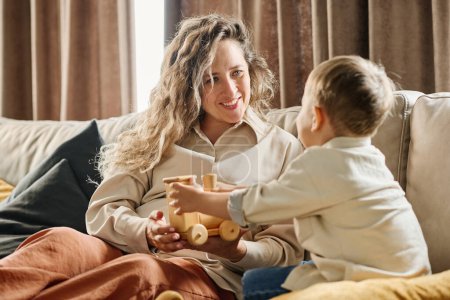 Photo for Happy young mother looking at her little son with smile while showing him new wooden toy during rest on couch in living room - Royalty Free Image