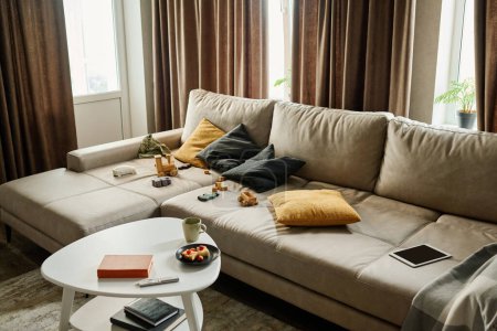 Foto de Parte de la amplia sala de estar en apartamento moderno con cómodo sofá de cuero gris con juguetes y cojines y pequeña mesa de pie en frente - Imagen libre de derechos