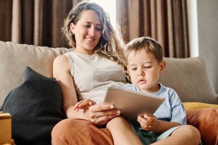 Téléchargez les photos : Petit garçon curieux regardant attentivement l'écran de la tablette tout en regardant un film pour enfants ou des dessins animés avec sa mère à la maison - en image libre de droit