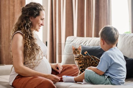 Téléchargez les photos : Heureuse jeune femme enceinte regardant son mignon petit fils jouer avec chat de race bengale tout en se relaxant sur le canapé dans le salon - en image libre de droit