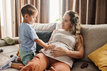 Foto de Curioso niño manteniendo la mano en el vientre de su madre embarazada sentada en el sofá mientras pasa tiempo libre con su lindo hijo pequeño en casa - Imagen libre de derechos