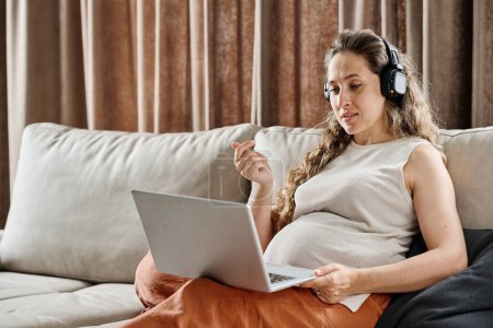 Photo for Young confident pregnant woman in headphones communicating with someone in video chat at home while looking at laptop screen - Royalty Free Image