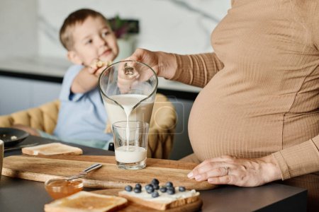 Téléchargez les photos : Gros plan de la jeune femme enceinte versant du lait frais dans du verre pour son mignon petit fils assis dans un fauteuil devant la table de cuisine - en image libre de droit