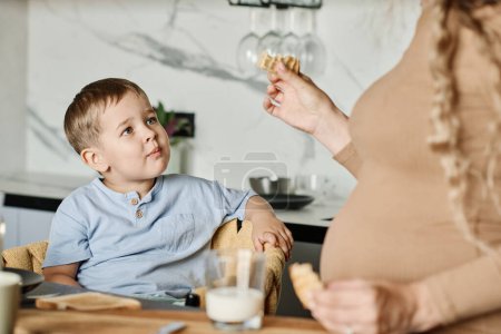 Foto de Lindo niño mirando a su madre embarazada con un pedazo de galleta casera en la mano mientras desayuna por la mesa de la cocina - Imagen libre de derechos