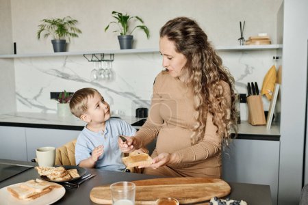 Foto de Mujer embarazada joven preparando sándwich con miel o mermelada para su adorable hijito mientras lo sostiene sobre la mesa de la cocina - Imagen libre de derechos