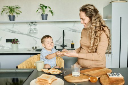Téléchargez les photos : Jeune mère prudente préparant un sandwich avec du miel ou de la confiture et le passant à son heureux petit fils assis dans un fauteuil près d'une table de cuisine - en image libre de droit