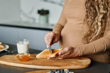 Téléchargez les photos : Mains de jeune femme enceinte avec couteau étalant du miel ou de la confiture sur une tranche de pain de blé frais pendant la préparation du petit déjeuner - en image libre de droit