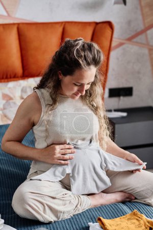 Téléchargez les photos : Jeune femme enceinte tenant un nouveau pull pour le futur bébé par le ventre et le regardant tout en étant assis sur un lit double dans la chambre - en image libre de droit