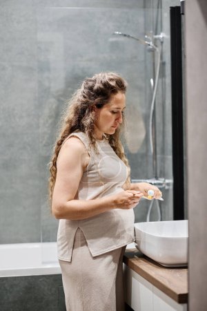 Foto de Mujer embarazada joven en casa usar apretando la pasta de dientes en el cepillo de dientes sobre el fregadero mientras cuida de sus dientes por la mañana - Imagen libre de derechos