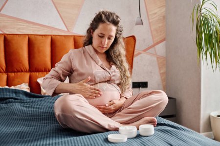 Foto de Mujer embarazada joven hidratando la piel de su vientre con crema hidratante mientras se sienta en la cama doble y se prepara para dormir - Imagen libre de derechos