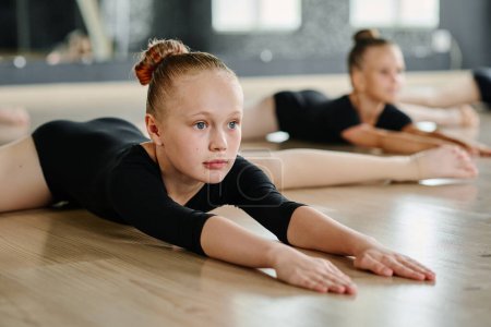 Téléchargez les photos : Fille flexible regardant vers l'avant tout en pliant et étirant les bras dans la pose de ficelle contre d'autres danseurs pendant la répétition ou le cours de ballet - en image libre de droit