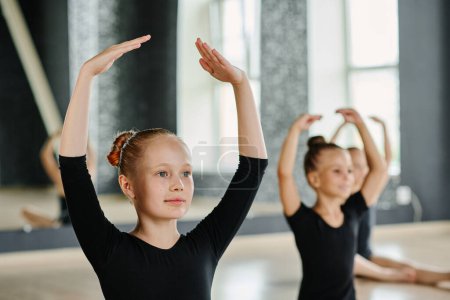 Téléchargez les photos : Jeune fille blonde en justaucorps noir gardant les bras levés au-dessus de la tête tout en pratiquant l'exercice chorégraphique à la leçon de danse - en image libre de droit
