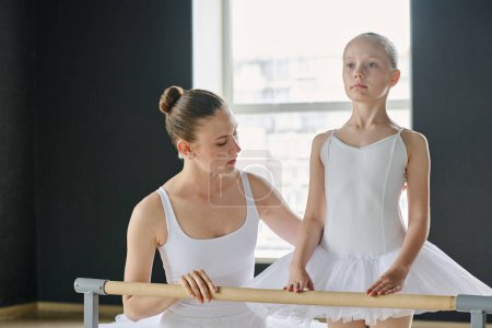 Foto de Joven instructora de ballet de pie junto a una joven aprendiz en tutú blanco durante la repetición en el aula y ayudándola con el ejercicio coreográfico - Imagen libre de derechos