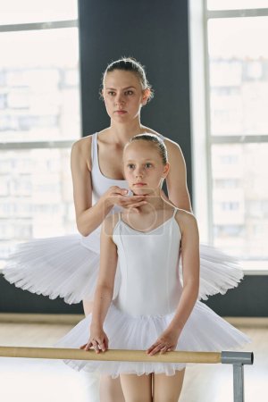 Foto de Joven profesora de ballet y su joven aprendiz en tutú blanco de pie junto a una barra de madera frente al espejo en el aula durante la repetición - Imagen libre de derechos