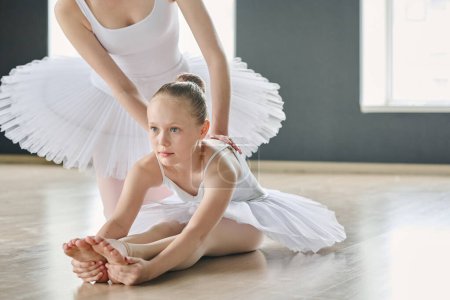 Photo for Cute youthful girl making effort while bending forwards over stretched legs while young ballet instructor helping her during exercise - Royalty Free Image