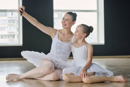 Foto de Alegre joven profesora de ballet y su joven aprendiz haciendo selfie en el teléfono inteligente mientras está sentada en el piso de la espaciosa sala de baile - Imagen libre de derechos