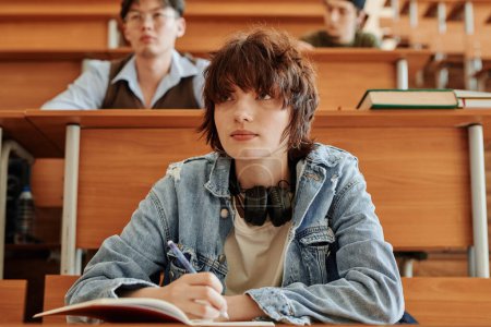 Photo for Cute teenage girl in denim jacket making notes while sitting by desk against her classmates and listening to teacher at lecture - Royalty Free Image