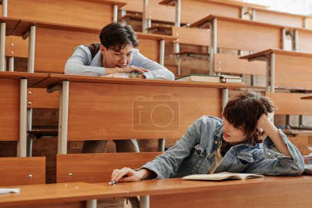 Foto de Dos jóvenes estudiantes interracial en ropa casual sentados junto a escritorios en el auditorio en el descanso, relajándose y charlando - Imagen libre de derechos