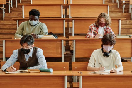 Groupe d'étudiants adolescents dans des masques de protection effectuant des tâches individuelles tout en étant assis à de longs bureaux dans la salle de conférence au collège