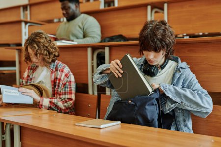 Téléchargez les photos : Adolescente brune en tenue décontractée mettre le livre dans le sac à dos après la leçon tout en étant assis par bureau dans l'auditorium contre ses camarades de classe - en image libre de droit