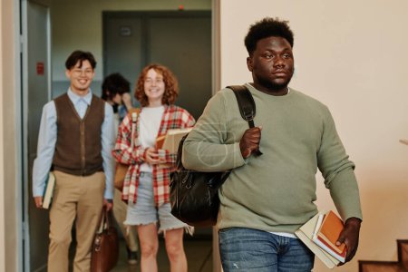 Téléchargez les photos : Jeune étudiant afro-américain en pull occasionnel gris entrant salle de conférence contre heureux couple interculturel debout dans la porte ouverte - en image libre de droit