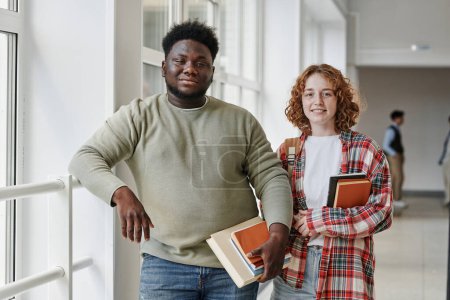 Youthful interracial classmates or couple with books standing in corridor of college or university after lessons and looking at camera