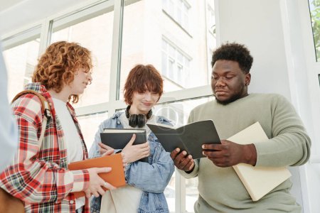 Téléchargez les photos : Afro-Américain gars et deux adolescentes regardant à travers les notes de cours dans son copybook tout en se tenant dans le couloir de l'université à la pause - en image libre de droit