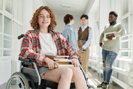 Photo for Smiling teenage girl with physical disability in casualwear sitting in wheelchair in college corridor against group of classmates - Royalty Free Image