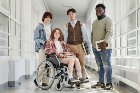 Foto de Grupo de estudiantes adolescentes multiculturales en ropa casual de pie en el pasillo de la universidad, mientras que uno de ellos sentado en silla de ruedas - Imagen libre de derechos