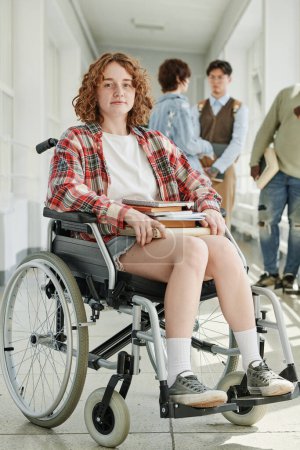 Foto de Linda estudiante adolescente en silla de ruedas sentada frente a la cámara en el pasillo de la universidad contra el grupo de sus compañeros de clase hablando en el descanso - Imagen libre de derechos