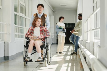 Téléchargez les photos : Heureux adolescent asiatique poussant sa petite amie en fauteuil roulant le long du couloir de l'université tout en avançant vers la salle de classe, bibliothèque ou cafétéria - en image libre de droit