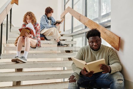 Téléchargez les photos : Jeune étudiant afro-américain et ses deux camarades de classe féminines lisant des livres assis sur un escalier dans le couloir de l'université à la pause - en image libre de droit