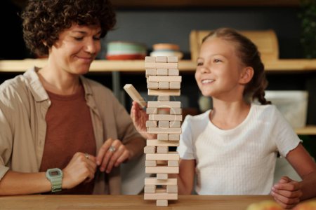 Photo for Cute girl with wooden block looking at high stack or tower standing on table while building it and her mother sitting next to her - Royalty Free Image