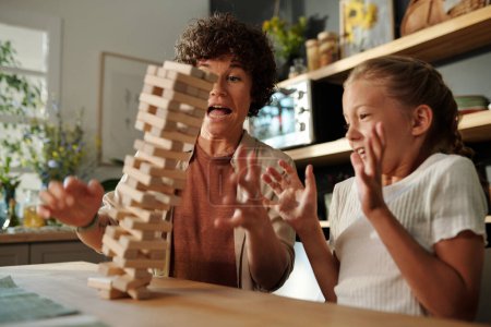 Téléchargez les photos : Jeune femme et sa jeune fille essayant de tenir sur la tour tombée construite de blocs de bois tout en étant assis à table pendant le jeu - en image libre de droit