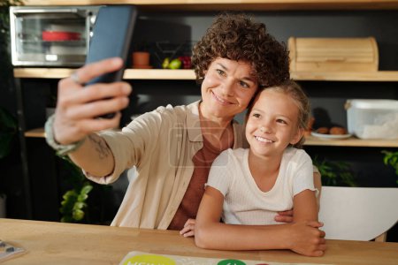 Photo for Happy young family of mother and daughter with smartphone making selfie while sitting by table at home and playing leisure game - Royalty Free Image