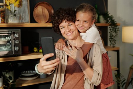 Photo for Young smiling mother with smartphone taking selfie with happy youthful daughter embracing her while standing behind at home - Royalty Free Image