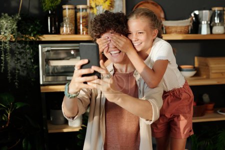 Foto de Linda chica joven cubriendo los ojos de su madre con el teléfono inteligente por las manos mientras se divierten durante selfie contra estantes con utensilios de cocina - Imagen libre de derechos