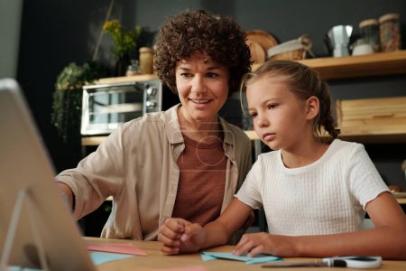 Téléchargez les photos : Jeune femme brune pointant vers l'écran de la tablette tout en étant assis à côté de sa fille et en l'aidant avec l'origami à loisir - en image libre de droit
