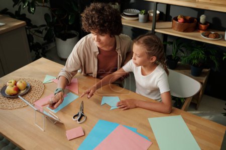 Foto de Mujer joven y su hija apuntando a la pantalla de la tableta durante el video en línea sobre el arte del origami mientras se sienta junto a la mesa en la cocina - Imagen libre de derechos