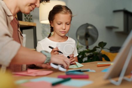 Téléchargez les photos : Jolie fille regardant sa mère pliant papier tout en créant origami à loisir après avoir regardé cours en ligne ou masterclass - en image libre de droit
