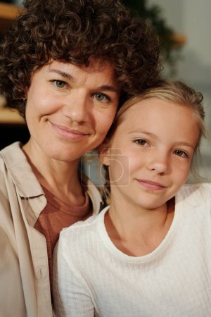 Photo for Happy young woman with short dark curly hair and her youthful daughter in white t-shirt looking at camera while sitting next to each other - Royalty Free Image