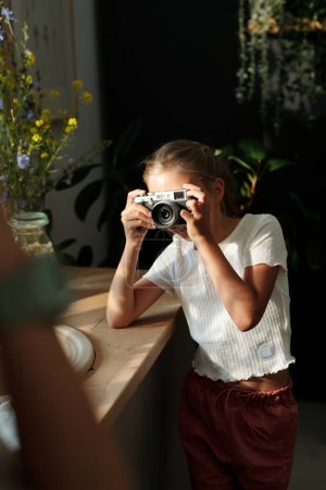 Téléchargez les photos : Jeune fille mignonne avec appareil photo rétro par son visage prenant une photo de sa mère ou de quelqu'un d'autre debout devant dans l'environnement de la maison - en image libre de droit