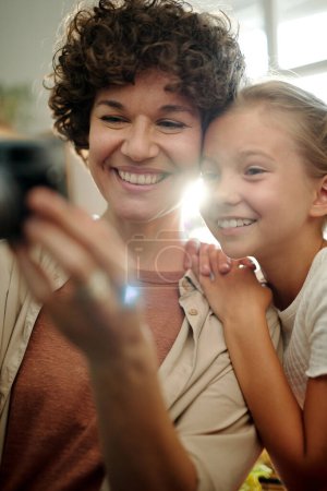 Photo for Cheerful young brunette woman with retro photocamera taking photo of herself and her cute youthful daughter standing near by - Royalty Free Image