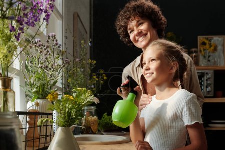 Foto de Feliz joven mujer de pie detrás de su linda hija refrescando flores mientras presiona pulverizador de botella de plástico verde con agua - Imagen libre de derechos
