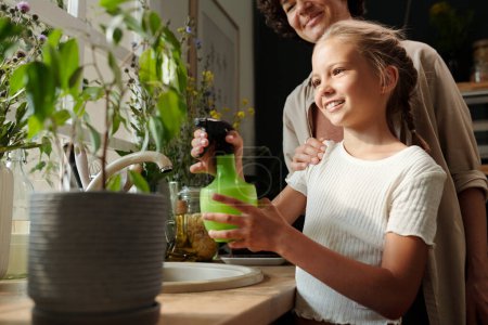 Téléchargez les photos : Jeune fille blonde mignonne avec bouteille en plastique vert rafraîchissant fleurs sauvages dans un vase tout en se tenant devant le pot de fleurs dans la cuisine - en image libre de droit