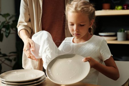 Téléchargez les photos : Petite fille mignonne en t-shirt blanc tenant un chiffon de coton tout en essuyant des assiettes propres après le lavage et en aidant sa mère à combattre la maison - en image libre de droit
