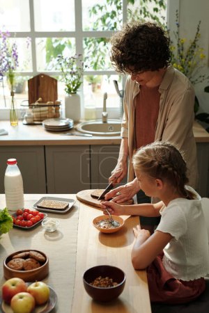 Foto de Mujer joven cortando pan de centeno fresco para sándwiches mientras prepara el desayuno junto a su hija joven comiendo muesli - Imagen libre de derechos